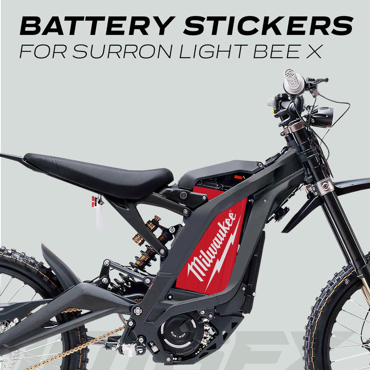 Sur-Ron Light Bee X with red battery sticker, displaying seat, rear shock, chain, and front fork against gray background. Highlighting the battery sticker as the focal point.