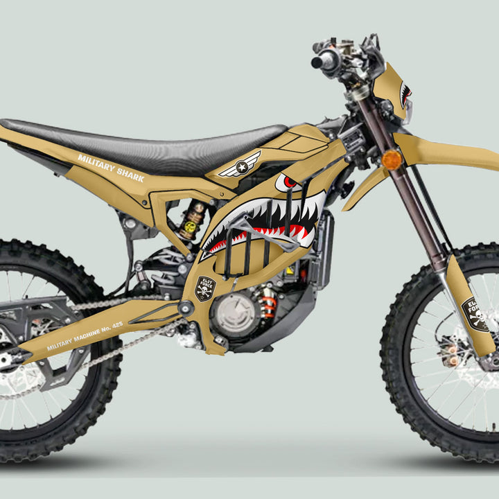 Side-view of a Surron Ultra Bee with Military Shark Sand camouflage graphics, featuring knobby tires, shark-mouth styling, and “MILITARY SHARK” stenciled text.